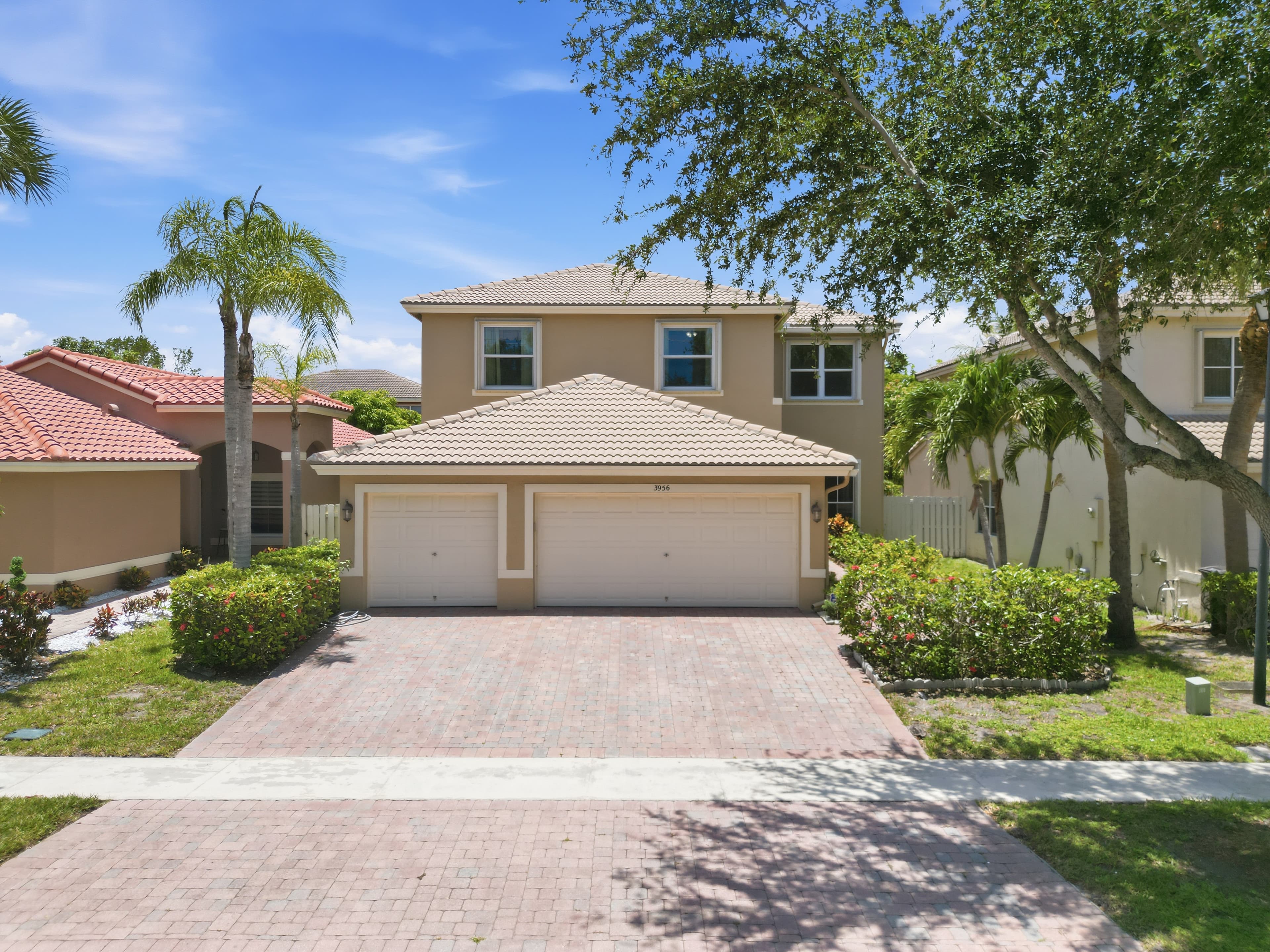 Tan two-story home with three-car garage, paver drive, and palms